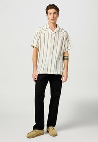 Camicia a maniche corte di colore crema con strisce nere verticali, chiusura a bottoni e vestibilità rilassata, abbinata a pantaloni neri e scarpe beige chiare.