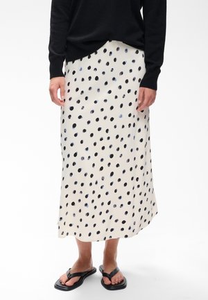 Personne portant un pull noir, une jupe midi crème à pois noirs et des sandales noires, debout sur un fond blanc uni.