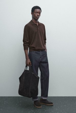 Man wearing brown long-sleeve polo, dark jeans, and dark shoes, holding large black tote bag, standing against plain light background.