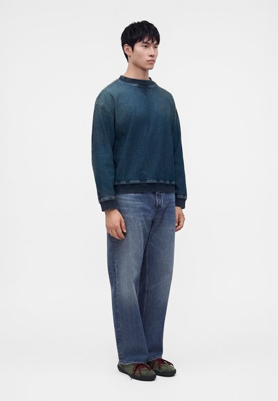 Jeune homme debout portant un sweat-shirt bleu foncé délavé, un jean bleu ample et des chaussures vert foncé avec des lacets rouges sur fond blanc.