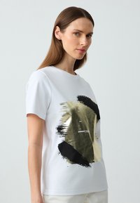 T-shirt bianca a maniche corte con un grande design astratto di pennellate oro e nero sul davanti, realizzato in morbido tessuto di cotone.