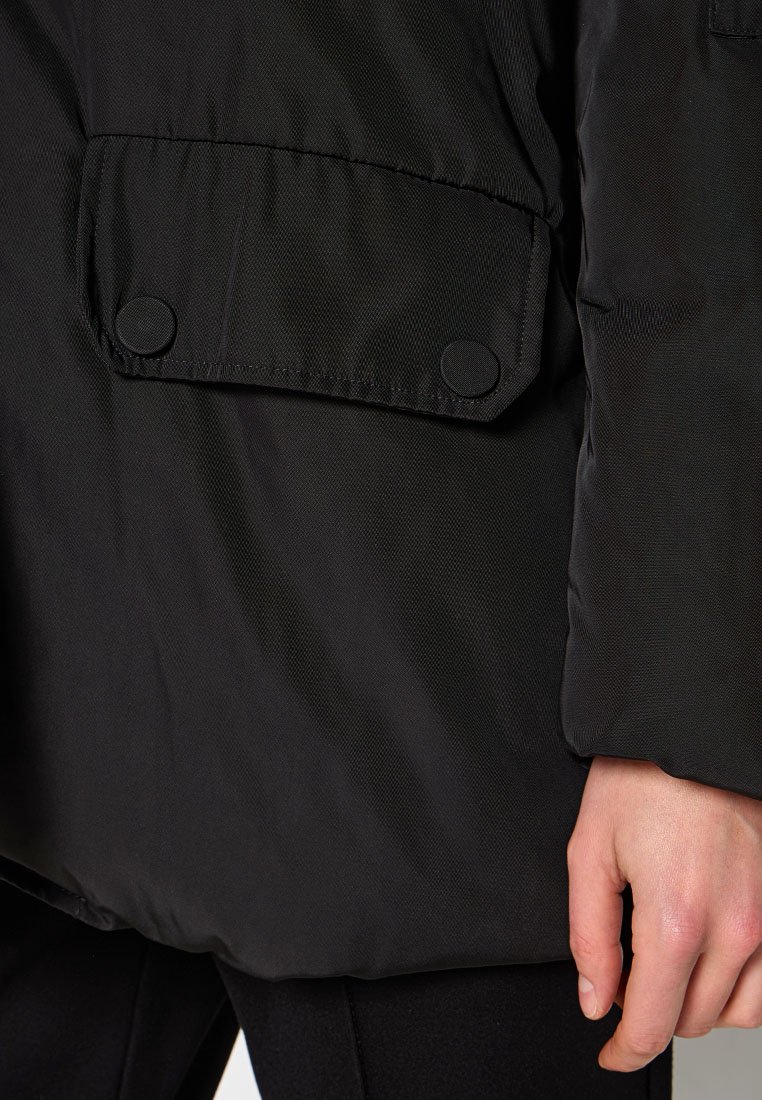 Schwarze Nylonjacke mit strukturiertem Finish, ausgestattet mit einer großen, knöpfbaren Tasche und einem lockeren Schnitt. Der Ärmel reicht bis zur Mitte des Unterarms.