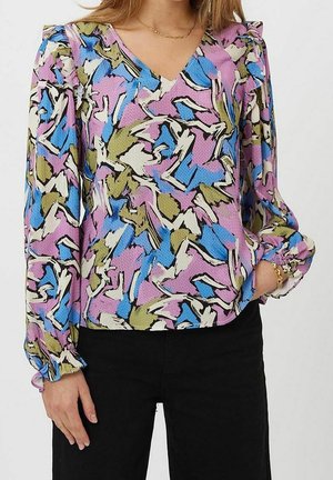 Bluzka z długim rękawem i dekoltem w serek z abstrakcyjnym kwiatowym wzorem w kolorach niebieskim, różowym, zielonym i białym, zestawiona z czarnymi spodniami i złotą biżuterią.