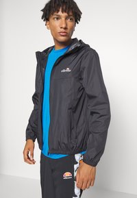 Schwarze Zip-Jacke mit Kapuze, elastischen Bündchen und Saum. Tragen über einem leuchtend blauen Shirt, kombiniert mit gemusterten Hosen mit orangefarbenen Akzenten.