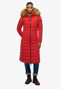 Rød puffjakke med hætte trimmed med pels, lang design, quiltet tekstur, fuld frontlynlås og sidelommer. Brune snøreboots er på.