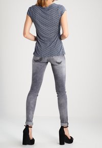 Modrý vzorovaný top s krátkým rukávem, šedé skinny džíny a černé vysoké podpatky. Oblečení má přiléhavý střih a hladkou texturu.