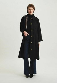 Manteau noir avec un grand col et des boutons dorés, superposé à un col roulé noir et un jean noir à jambes larges. Il présente un accent de chaîne décorative.