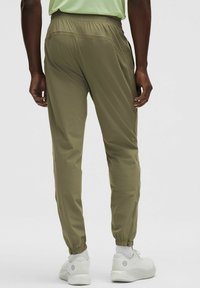 Olivegrüne tapered Jogginghosen mit elastischem Bund, Reißverschlusstaschen und elastischen Bündchen, kombiniert mit weißen Sneakers vor einem neutralen Hintergrund.