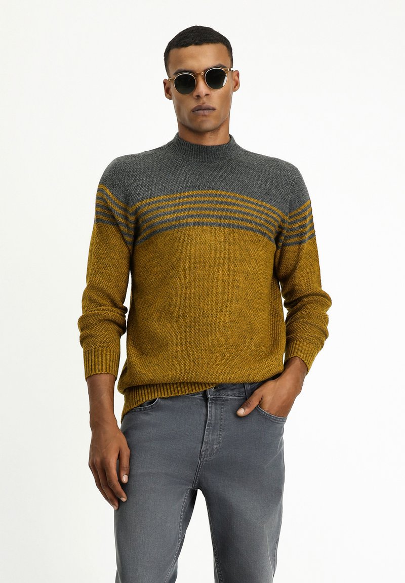 Gestrickter Pullover in Gelb- und Grautönen mit horizontalen Streifen, mit Rundhalsausschnitt und gerippten Bündchen. Modell trägt graue Jeans und Sonnenbrille.