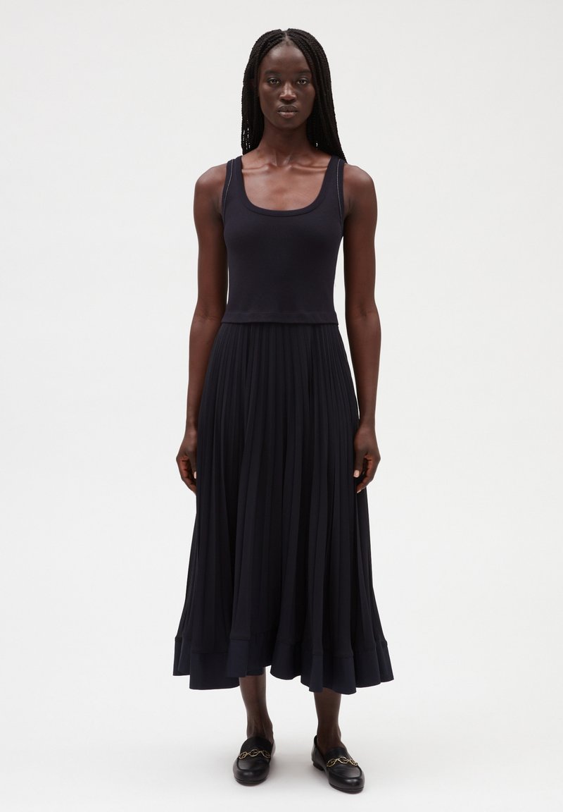 Vestido maxi azul marino con falda plisada y parte superior ajustada. Tejido suave, escote redondo y dobladillo acampanado. Zapatos negros de-slip-on con detalles de cadena.