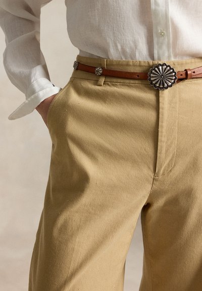 Άτομο που φοράει μπεζ παντελόνι με καφέ δερμάτινη ζώνη με ασημένια λουλουδένια κουμπώματα και κρεμ πουκάμισο με κουμπιά, βαλμένο μέσα.