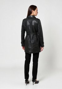 Zwarte leren trenchcoat met ceintuur in de taille, schouderepauletten en lange mouwen, gecombineerd met zwarte strakke broek en hoge hakken.