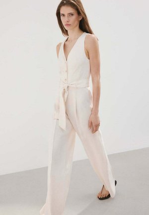 Frau geht, trägt einen ärmellosen cremefarbenen Overall mit Knöpfen und gebundenem Taillengürtel, kombiniert mit schwarzen Sandalen.