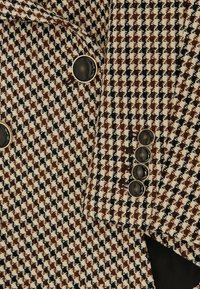 Brown, black, and white houndstooth patterned blazer with black buttons edged in silver on sleeve and front.