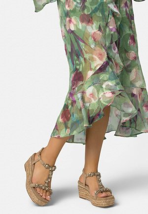 Pies de mujer con sandalias cuña beige adornadas con joyas florales, vistiendo un vestido verde con estampado floral, de vuelo, con bajo con volantes y una abertura.