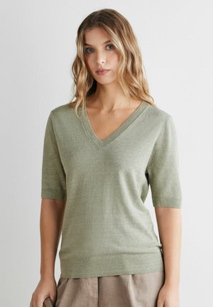 Kvinde med bølget blondt hår iført en lysegrøn V-hals kortærmet sweater og beige bukser mod en ensfarvet baggrund.