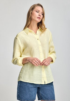 Camisa de manga larga color amarillo claro, de corte holgado, hecha de lino texturizado, con botones en la parte frontal y un pequeño logotipo en el lado izquierdo del pecho.