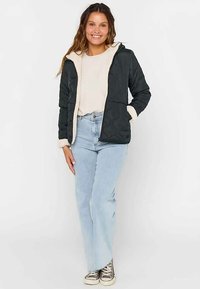 Zwarte doorgestikte jas met een crèmekleurige fleece gevoerde capuchon, rits en zakken, gedragen over een crèmekleurige trui en lichtblauwe spijkerbroek.