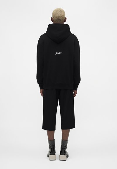 Oseba s kratkimi blond lasmi, ki nosi črno kapucar s belim napisom na hrbtu, črne pohodne hlače in črne gležnjarje s kvadratnimi petami.