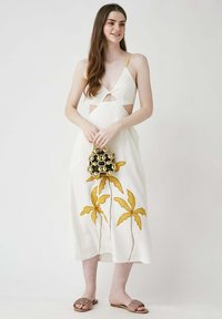 Weißes Kleid mit Ausschnitt-Details, gelben Akzenten und gelber Blumenstickerei. Das Modell hält eine schwarze runde Handtasche mit goldenen Verzierungen.