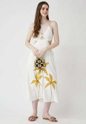 Weißes Kleid mit Ausschnitt-Details, gelben Akzenten und gelber Blumenstickerei. Das Modell hält eine schwarze runde Handtasche mit goldenen Verzierungen.