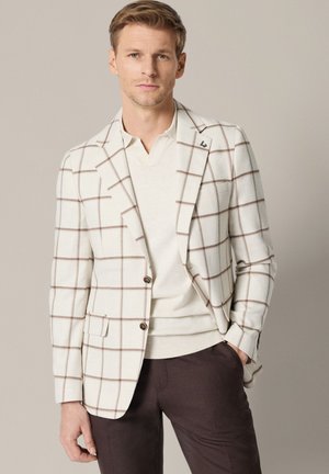 Mężczyzna w białej marynarce w brązową kratę, kremowym swetrze z dekoltem w serek i ciemnobrązowych spodniach, stojący na jednolitym tle.