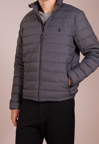 Grå puffjacka med kviltad söm, hög krage och dragkedja fram. Har sidofickor och minimal logotyp.