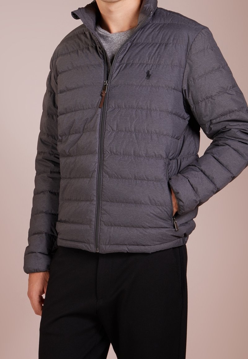 Grå puffjacka med kviltad söm, hög krage och dragkedja fram. Har sidofickor och minimal logotyp.