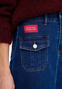 Džínsy vyrobené z tmavomodrej látky, s kontrastným žltým prešívaním a červenou nálepkou s bielym textom na zadnej vrecku.