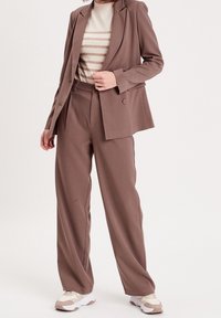 Blazer marron sur mesure avec un pantalon large assorti et un pull rayé beige en dessous. Caractéristiques d'un tissu lisse et de détails de boutons.