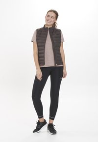 Mjuk, mörkbrun väst över en ljusrosa skjorta, kombinerad med svarta leggings och svarta träningsskor. Slät textur och figurnära design.