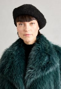 Fauxfurmantel in tiefem Teal mit einer weichen, plüschigen Textur; über einem schwarzen Rollkragenpullover getragen, kombiniert mit einem strukturierten schwarzen Baskenmütze.