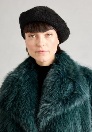 Sünteetilisest karusnahast sügava teal värvi mantel pehme, plush tekstuuriga; kantud musta kõrge kaelusega särgi peal, koos tekstuuriga musta beretiga.