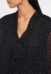 Blouse noire transparente à pois, avec un col à nouer et des manches courtes. Présente des taches blanches, une poche poitrine et une coupe ample et décontractée.