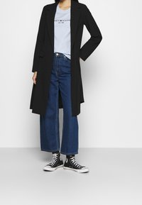 Musta pitkä takki vaaleansinisellä T-paidalla, yhdistettynä leveisiin farkkuihin ja mustiin korkeakantaisiin lenkkareihin. Silkkinen kangas ja rento muotoilu.