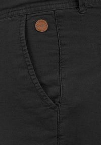 Pantalones negros de tejido texturizado, con un bolsillo lateral y una etiqueta de cuero circular con una marca en relieve.