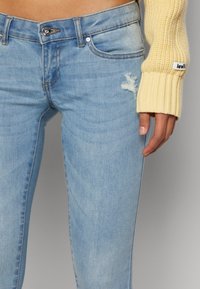 Vaqueros de mezclilla azul claro con un parche desgastado, detalle de botón plateado y costuras en contraste. Combinados con un suéter amarillo acanalado.