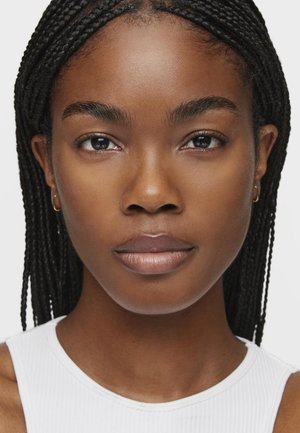 Primer plano de una persona con cabello trenzado, tono de piel claro, vestida con una camiseta blanca de canalé. Sus características incluyen cejas definidas y pequeños pendientes de aro.