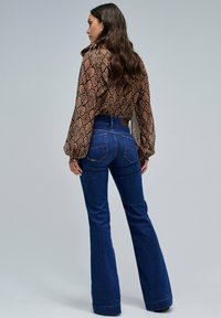 Blusa marrón con estampado de serpiente y mangas largas, combinada con jeans de campana azules. El conjunto presenta una cintura ajustada y detalles sutiles en los bolsillos traseros.