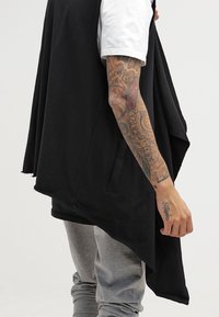 Person som bär en svart asymmetrisk ärmlös kappa över en vit skjorta och grå byxor, som visar en helt tatuerad högra arm och hand.