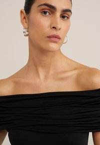 Top nero scollato con texture arricciata, realizzato in un tessuto liscio. Modello che indossa orecchini a cerchio in argento su uno sfondo neutro.