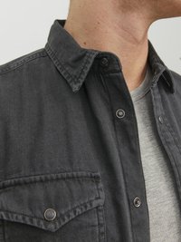 Camisa de un gris oscuro con textura suave, que presenta un cuello clásico, un bolsillo en el pecho y botones a presión, superpuesta a una camiseta básica de un gris claro.