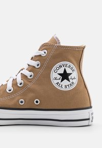 Brown canvas high-top sneaker with white laces, black rubber sole, and a round black-and-white logo patch featuring a star.