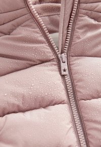 Gros plan d'une veste matelassée rose poussiéreuse avec des gouttes d'eau sur le tissu et une fermeture éclair en plastique partiellement fermée avec un tirette en forme d'étoile.