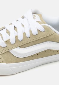 Beige suede sneaker with white rubber sole, featuring white laces, a white wave design, and perforations along the sides for ventilation.