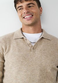 Beige gestrickter Polo-Pullover mit V-Ausschnitt und drei Knöpfen am Kragen. Über einem weißen T-Shirt getragen, das über eine dezente Textur und eine weiche Oberfläche verfügt.