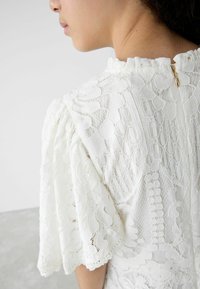 Witte kanten top met puffmouwen, ingewikkelde bloemen- en gaaspatronen, gerimpelde halslijn en een subtiele gouden rits aan de achterkant.