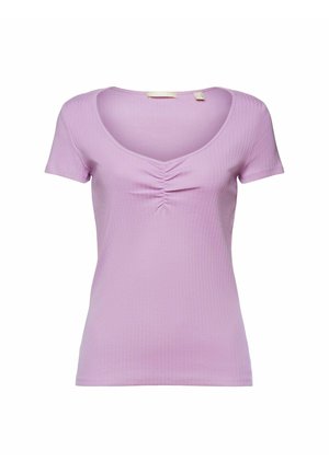 Kobieta ubrana w fioletowy top z krótkim rękawem zawiązany w supeł, czarne spodnie z wysokim stanem i czarne sandały, stojąca na tle żółtej ściany.