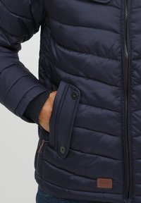 Navy padded jacket with horizontal stitching, featuring a zipped front, snap-button pocket, and ribbed cuffs. Light brown brand label detail.