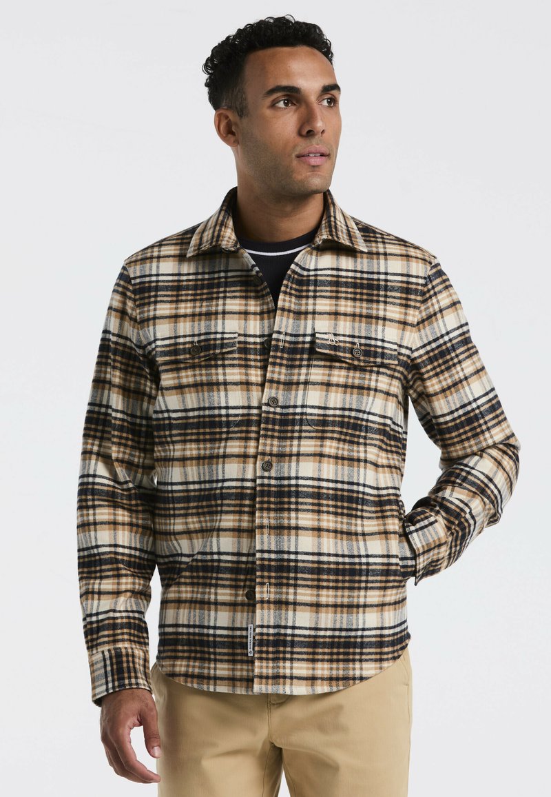 Man in een beige, zwart en witte geblokte overhemd met knopen en beige broek, kijkend naar rechts, met één hand in zijn broekzak tegen een effen achtergrond.